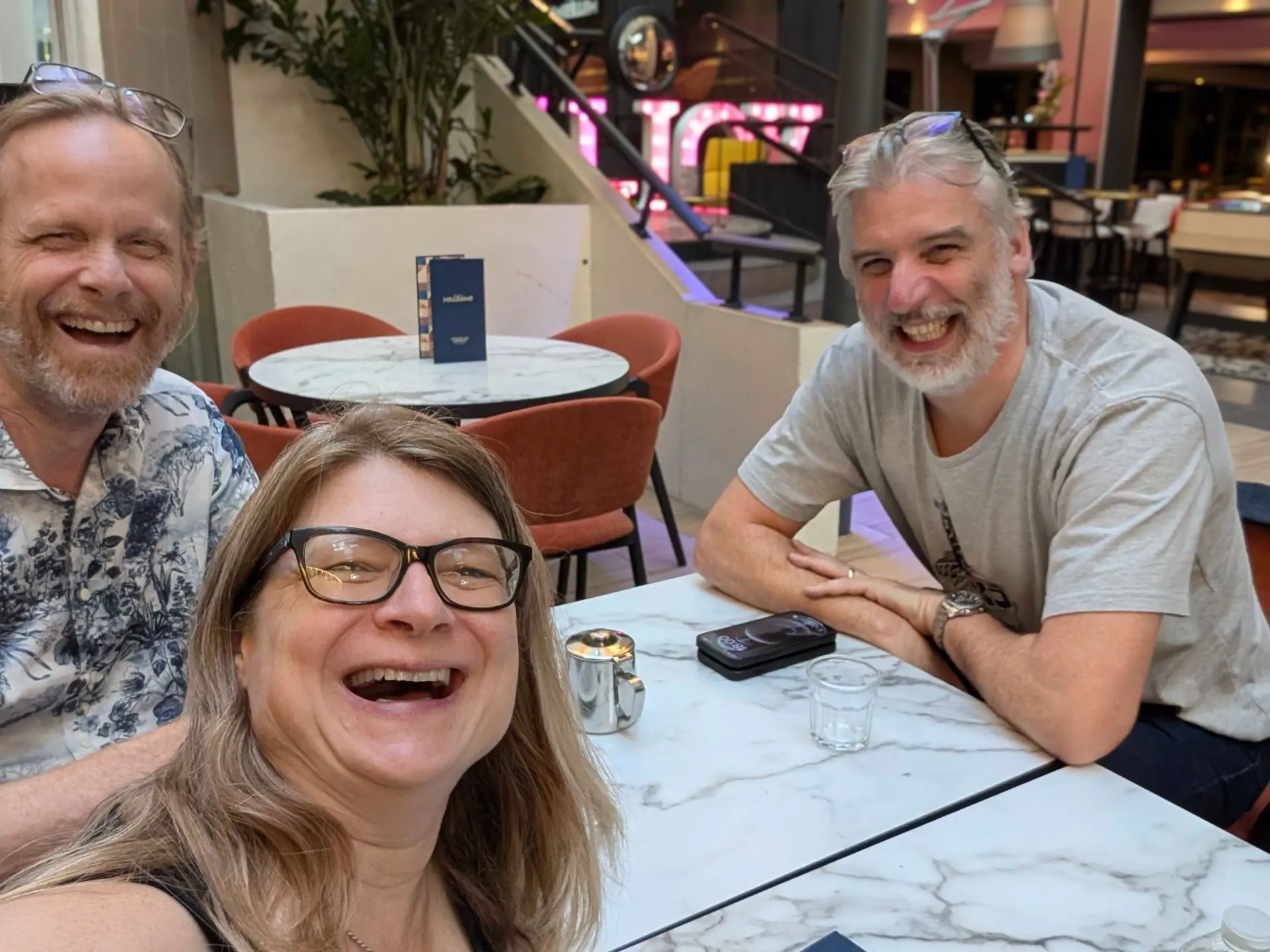 Three people smiling at a cafe table with marble tops and orange chairs, background features a staircase with pink neon signs and a plant. The group consists of two men and a woman, all with glasses on or near their heads, sitting and posing for a photo.